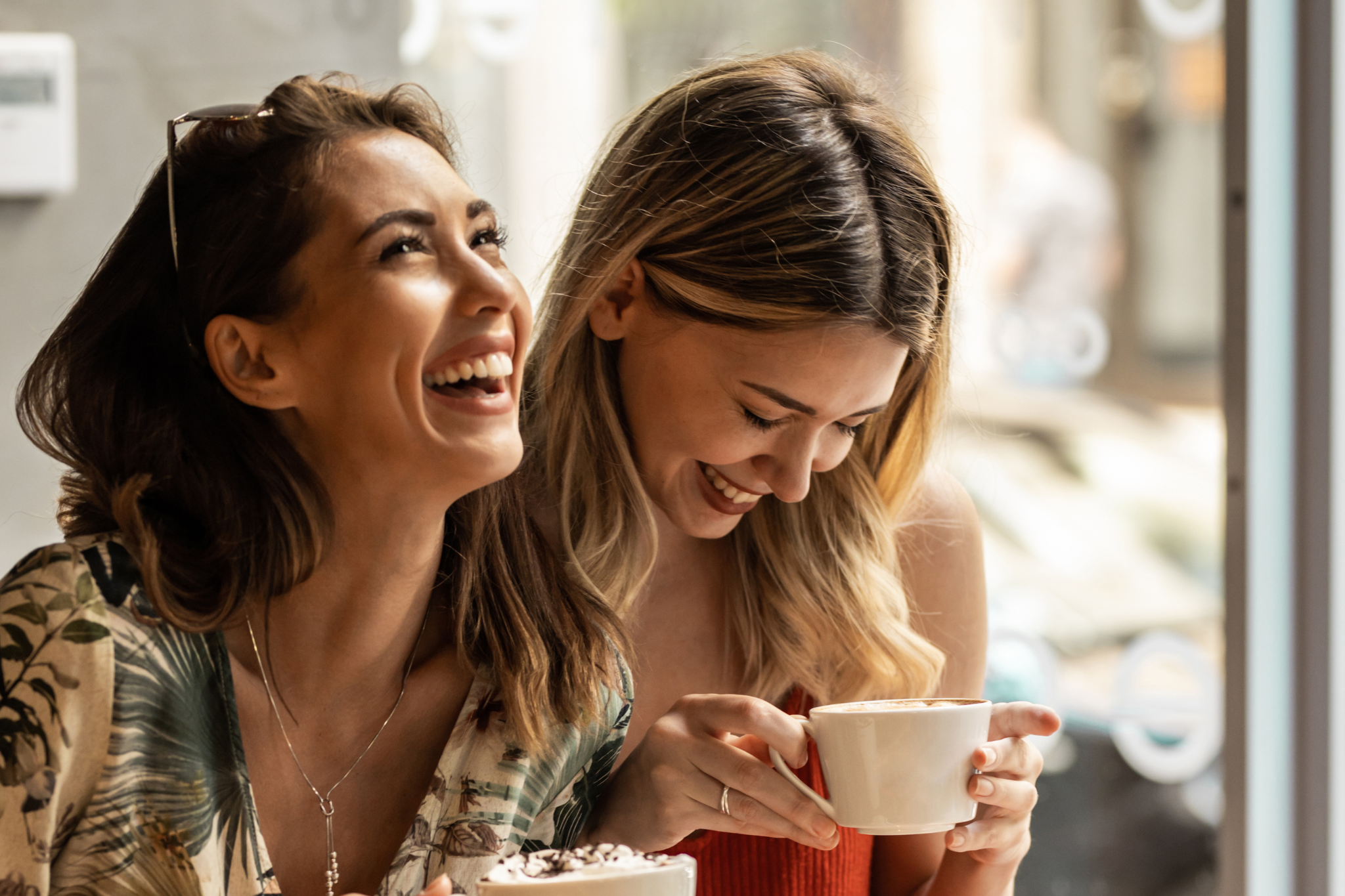 Zwei lachende Personen sitzen in einem Café und genießen Getränke. Eine Person hält eine Tasse, die andere ein Dessert. Beide wirken entspannt und fröhlich.