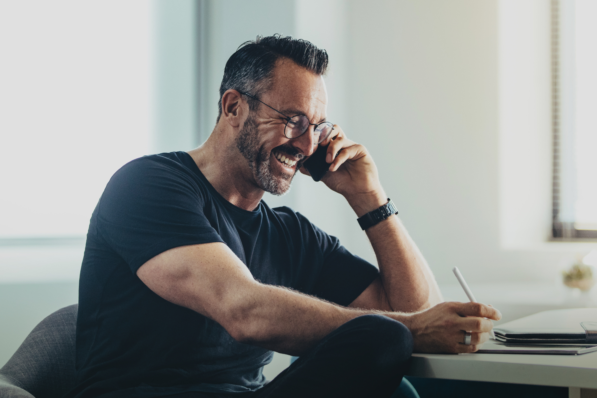 Lächelnder Mann mit Brille und Bart telefoniert im Büro und macht sich Notizen mit einem Stift. Er trägt ein schwarzes T-Shirt und sitzt an einem hellen Schreibtisch.