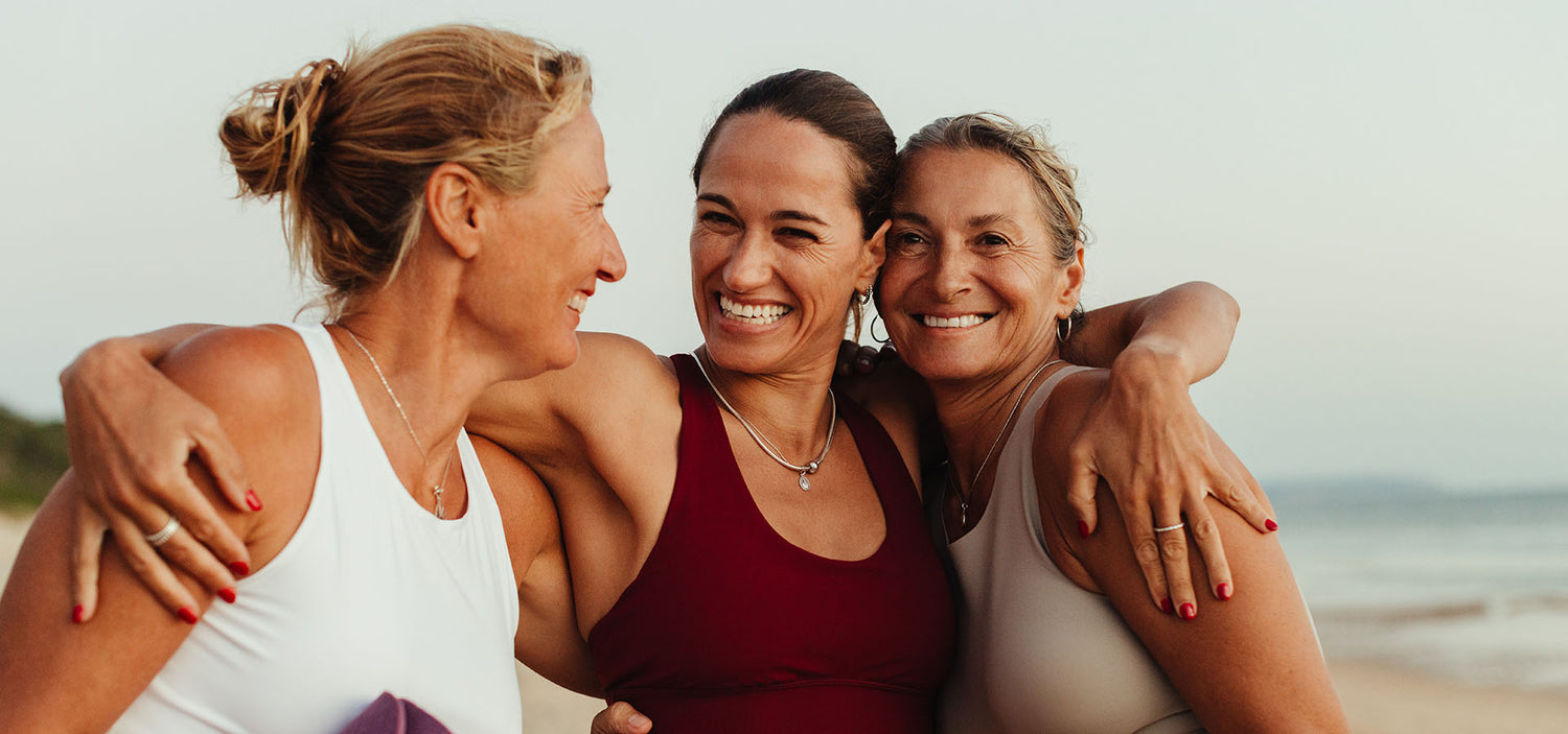 Drei sportliche Frauen umarmen sich lachend am Strand und strahlen Lebensfreude, Energie und Zusammenhalt aus.