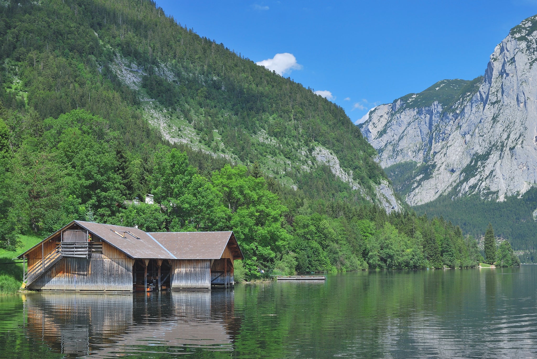 Idyllischer Bergsee mit Holzhütte am Ufer, grünen Wäldern und imposanten Felsen im Hintergrund bei blauem Himmel – Naturidylle und Ruhe in den Alpen.