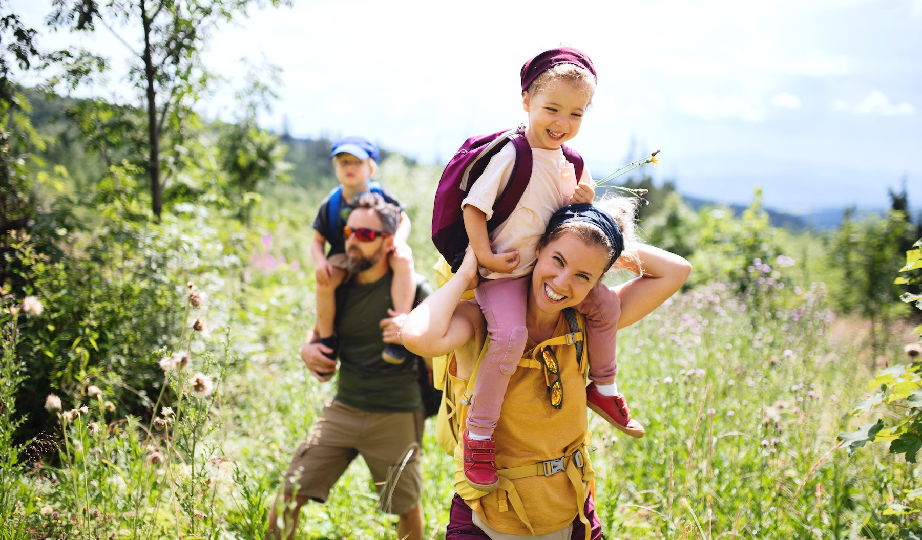 Glückliche Familie beim Wandern in den Bergen – Mutter und Vater tragen ihre Kinder auf den Schultern durch eine blühende Wiese, Natururlaub und Familienzeit in den Alpen.