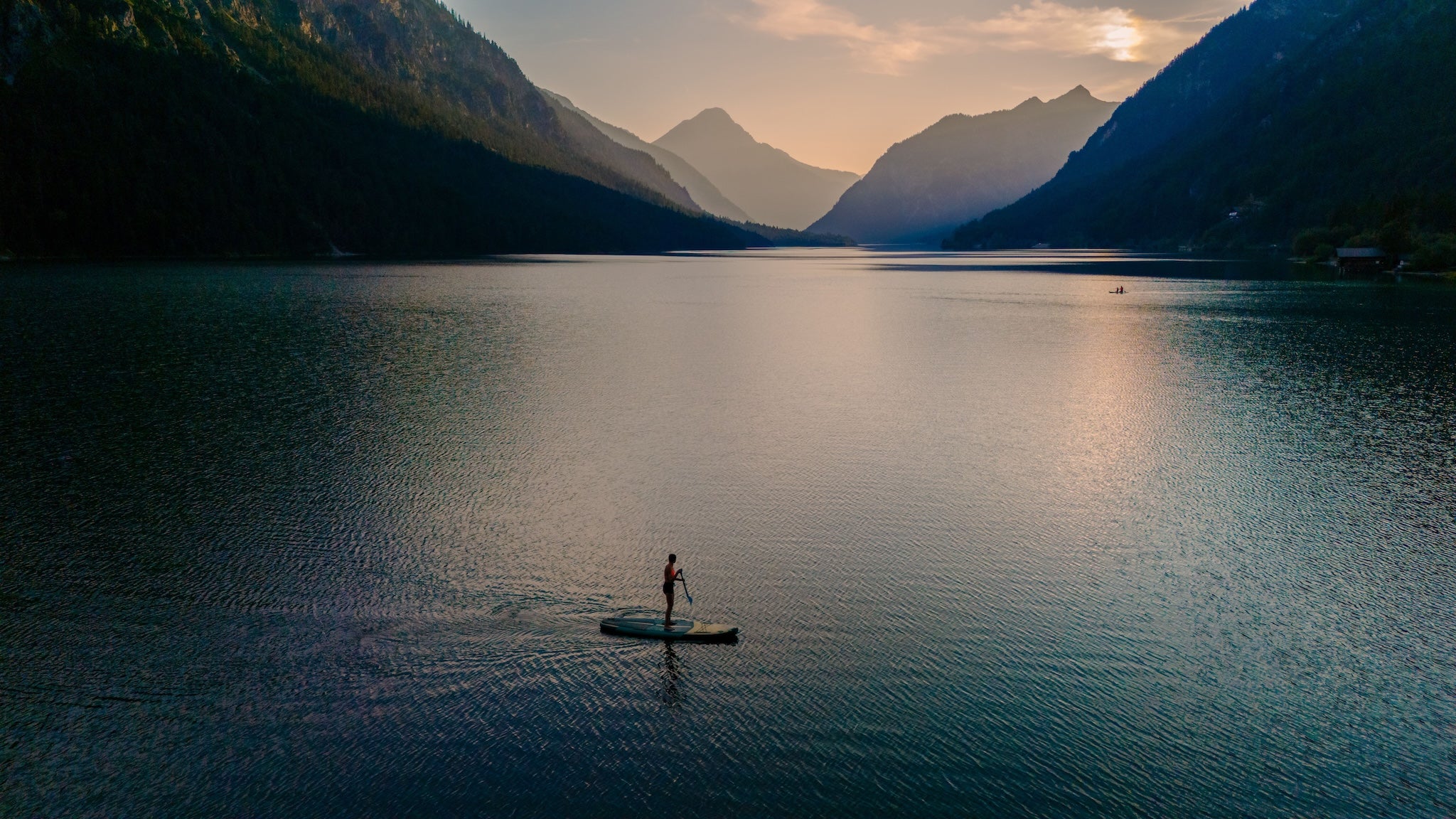 Stand-up-Paddler auf einem ruhigen Bergsee bei Sonnenuntergang, umgeben von Alpenlandschaft – Symbol für Natur, Balance und gesunden Lifestyle.